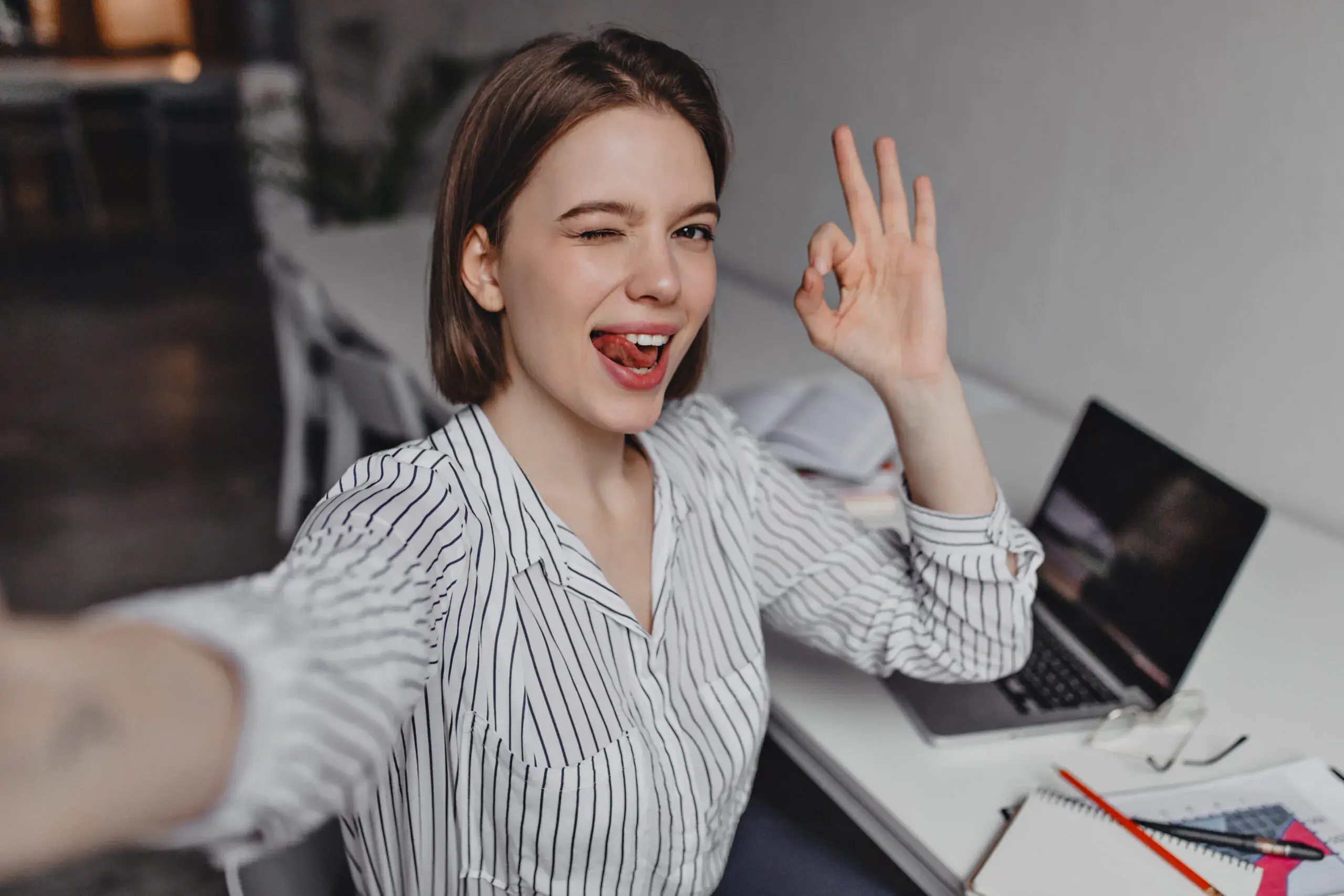 mischievous-woman-takes-selfie-in-office-office-worker-in-striped-shirt-shows-sign-ok-on-background-of-laptop-scaled