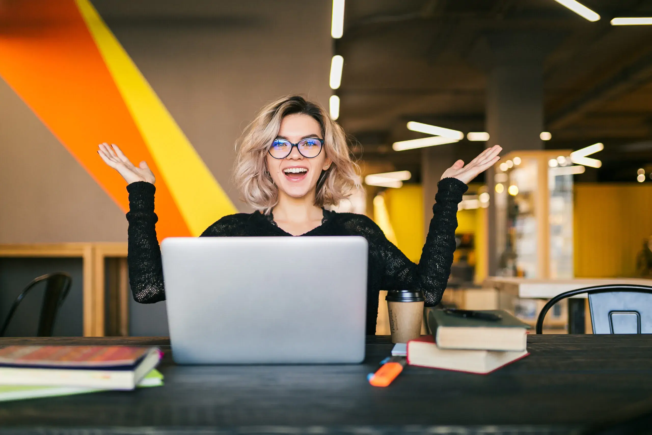 funny-happy-excited-young-female-sitting-at-the-table-in-a-black-shirt-working-on-a-laptop-in-an-office-working-on-her-glasses-scaled